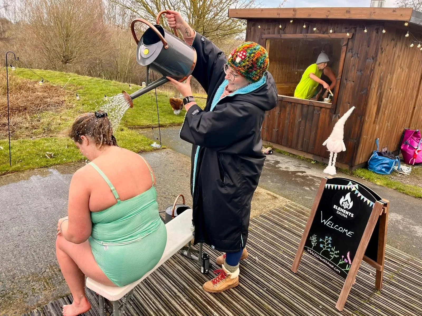 A swimmer being doused with a watering can of cold water on the jetty outside the sauna