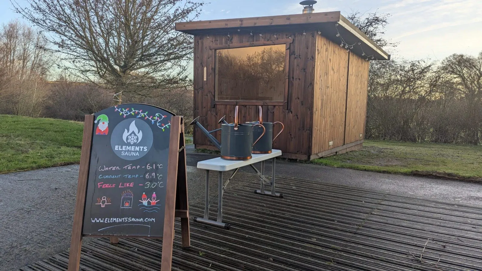 The sauna cabin in low golden light, with the Elements Sauna A-board and two watering cans on a bench outside