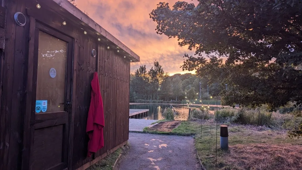 The sauna door under an orange sky, a red robe hanging beside it, fairy lights along the roof and the jetty beyond