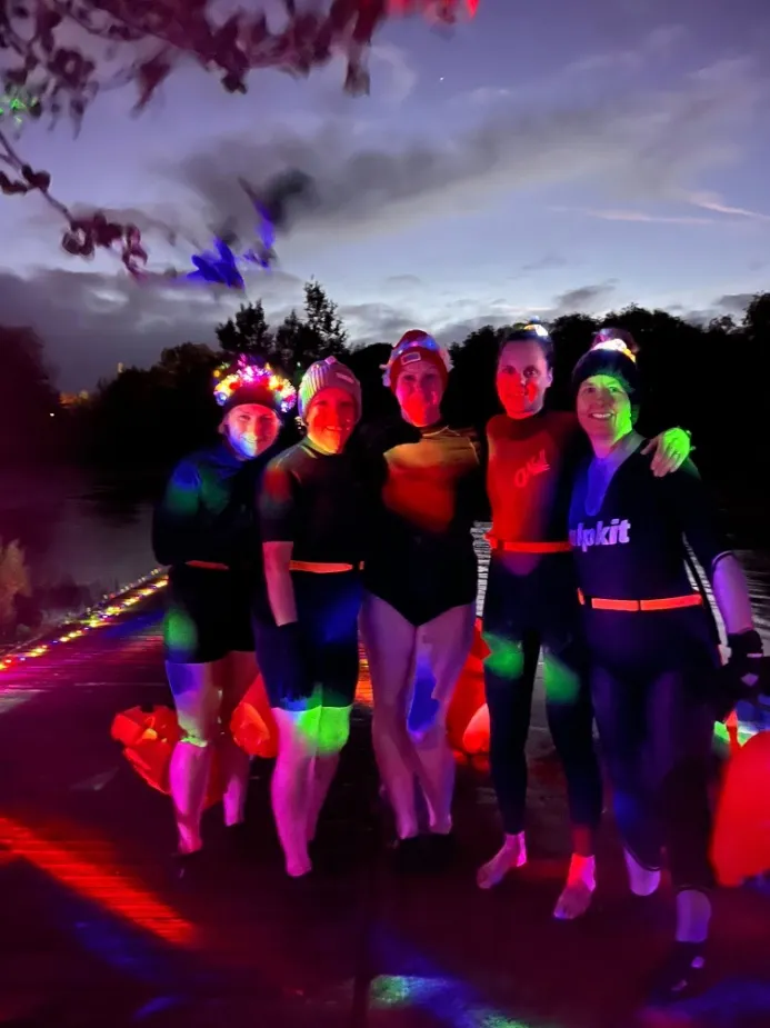 Five swimmers in light-up hats on the jetty at a Light-Up night, lit by coloured light