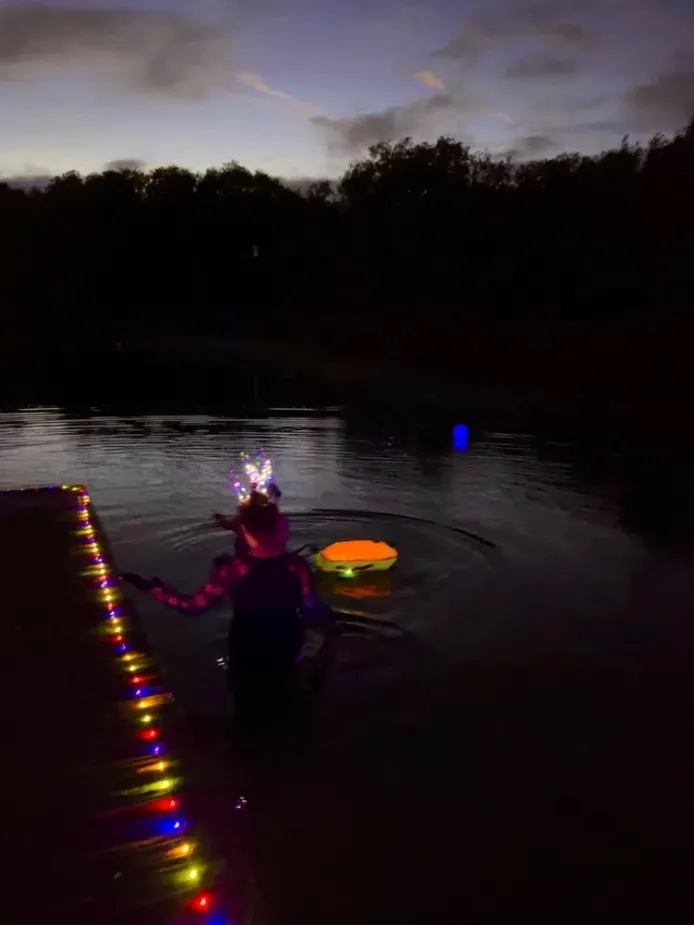 A swimmer with a glowing float stepping into the dark lake beside a lit-up jetty