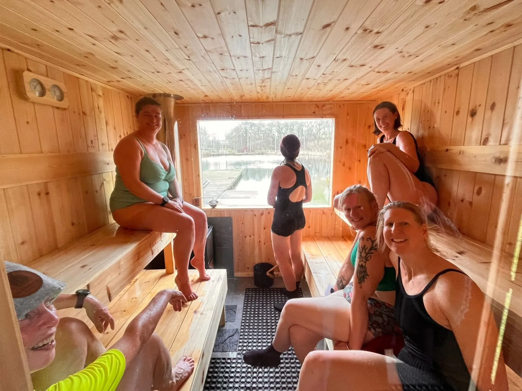 Six women laughing together on the sauna benches with the lake behind them