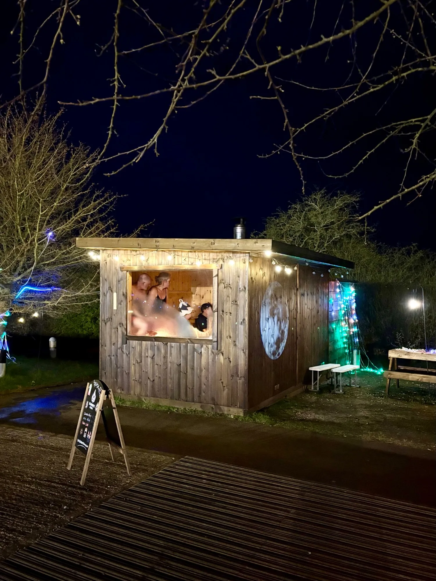 The sauna at night for a full moon session, lit with fairy lights and glowing from inside, with a moon painted on its side