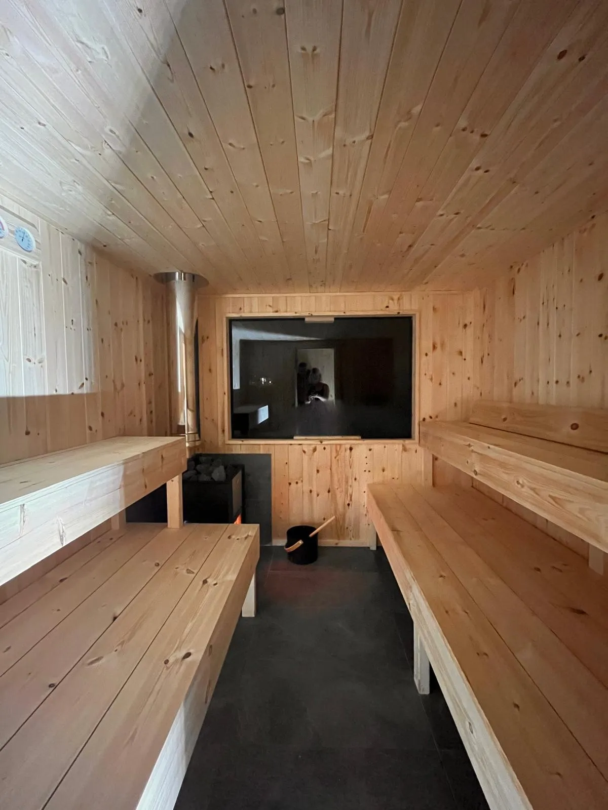 Inside the sauna: pale pine benches on two levels, the stove with its stones, a bucket and ladle and a large window