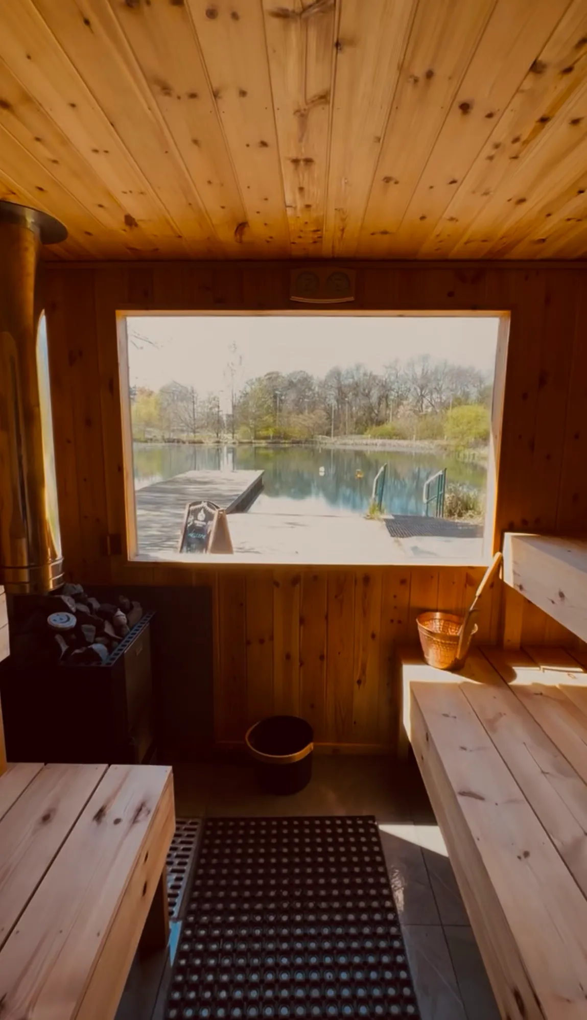 Sunlight in the sauna: pine ceiling, wood-fired stove with stones, copper bucket and the lake through the window