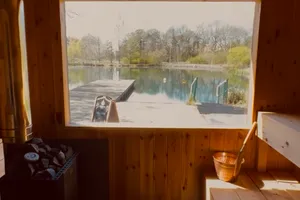 Sunlight in the sauna: pine ceiling, wood-fired stove with stones, copper bucket and the lake through the window