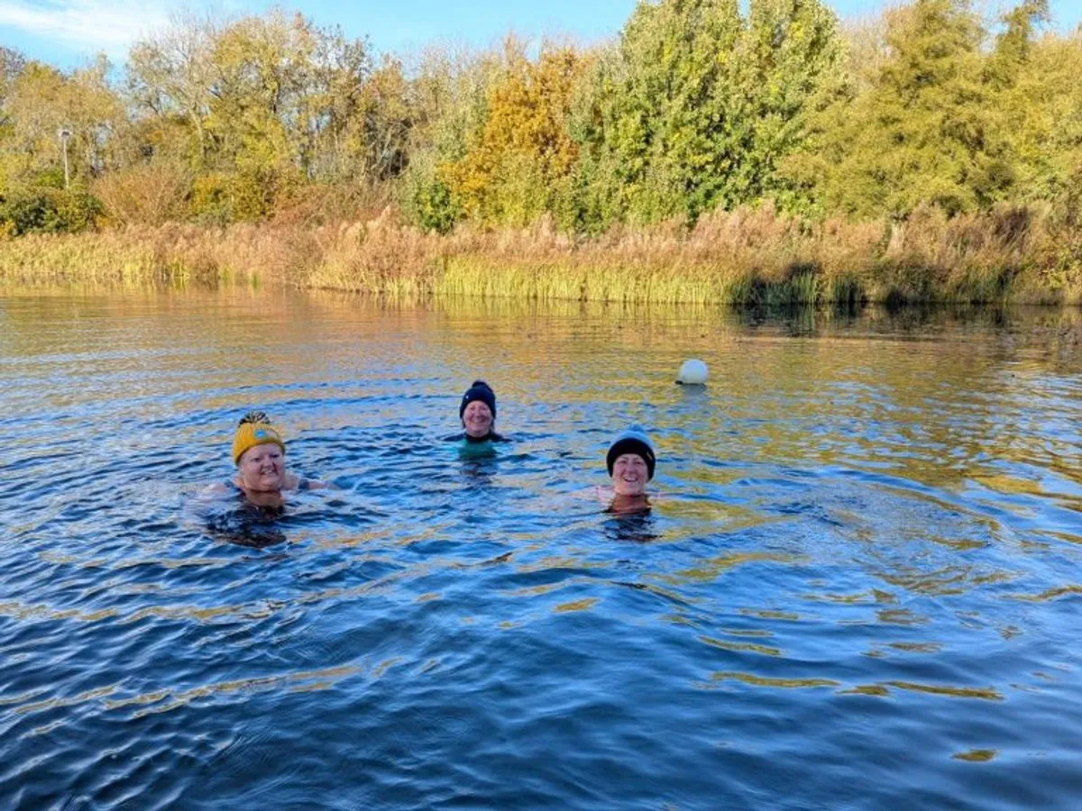 Three wild swimmers in the spring-fed lake at TCR Hub, Barnard Castle, with autumn trees behind